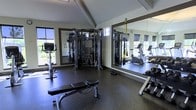 This single-floor fitness facility offers a thoughtfully designed workout environment suited for a wide range of exercise routines. The room features a striking vaulted ceiling that lends an open, airy quality to the space, while large windows along the walls allow natural light to fill the interior, creating an energizing atmosphere throughout the day.

The flooring consists of durable rubber matting, providing both safety and stability during workouts. A large wall-mounted mirror spans a significant portion of one wall, visually expanding the space and allowing users to monitor their form during exercise.

At the center of the room stands a multi-station functional cable machine, offering a comprehensive range of strength-training options. Complementing this is a padded adjustable workout bench and a dumbbell rack stocked with free weights of various sizes. Cardio equipment, including what appears to be a stationary bike and a treadmill, rounds out the facility's offerings. TRX suspension training straps are also mounted and ready for use, adding further versatility to available workout options. The overall ambiance is clean, modern, and functional.