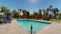 Located in Santa Clarita, California, this outdoor amenity area centers around a generously sized swimming pool with clear, blue-green water, designed to accommodate both leisurely swimming and relaxation. The pool is complemented by a separate spa, providing a dedicated space for unwinding. Surrounding the pool, a broad concrete sundeck offers ample room for lounge chairs and seating, ideal for enjoying the Southern California sunshine. The area is enclosed by a wrought iron security fence with gated access, ensuring a safe and private environment. Pergola structures provide shaded gathering spots throughout the deck, adding architectural interest and functional relief from the sun. The perimeter is richly landscaped with tall palm trees, mature deciduous trees, and lush greenery, creating a resort-like atmosphere that feels both secluded and inviting. Adjacent residential buildings with a dark exterior finish are visible, framing the space and contributing to the cohesive community setting. The overall ambiance is relaxed and park-like, with abundant natural surroundings that enhance the outdoor experience and offer a welcome retreat within the broader Santa Clarita community.