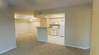 This single-floor space showcases an open-concept design that seamlessly connects the living area and kitchen, creating a cohesive and functional layout. The living area is finished with wall-to-wall carpet in a neutral beige tone, contributing to a warm and understated ambiance. A chandelier-style pendant light fixture is positioned overhead, adding a modest decorative touch to the space.

The kitchen is accessible via an open archway and features light wood cabinetry that extends to the upper walls, providing ample storage. White appliances, including a range, microwave, and refrigerator, are neatly integrated into the layout. A countertop peninsula with a granite-style surface serves as a natural divider between the kitchen and the living area, offering additional workspace and the potential for casual seating.

The overall color palette remains consistent throughout, with beige walls and light-toned surfaces creating a clean, cohesive aesthetic. A ceiling vent is visibly integrated into the ceiling near the kitchen transition, indicating a central HVAC system. The tile flooring in the kitchen contrasts subtly with the carpeted living space, clearly delineating the two functional zones.