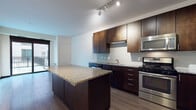 This well-appointed single-floor residence offers a thoughtfully designed layout with contemporary finishes throughout. The kitchen serves as the centerpiece of the living area, showcasing rich dark wood cabinetry, a distinctive penny-tile backsplash, and a full suite of stainless steel appliances including a gas range, built-in microwave, dishwasher, and refrigerator. A generously sized kitchen island topped with speckled granite provides additional prep space and casual seating, while track lighting overhead keeps the area bright and functional. The hardwood-style flooring in the kitchen and main living area adds warmth and cohesion to the open-concept space. The bedroom is carpeted for comfort and features large floor-to-ceiling windows and a sliding glass door that opens to a private outdoor balcony, allowing natural light to fill the room. A spacious walk-in closet with built-in wire shelving and hanging rods offers ample storage. The bathroom is clean and modern, finished with large-format gray tile surrounding a full soaking tub and shower combination, complemented by light tile flooring and simple, neutral walls. Overall, the space strikes a balance between modern urban design and comfortable everyday living.