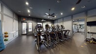 This fitness center is thoughtfully designed to accommodate a full range of workout needs within a single, open floor plan. The space is divided into distinct functional zones, each tailored to a specific type of training. The cardio area houses a substantial row of modern elliptical trainers and stair climbers equipped with individual digital display screens, allowing users to track performance metrics during their sessions. Treadmills and additional cardio equipment round out this section.

The strength training zone is equally well-appointed, featuring a multi-functional cable machine, a dedicated free weights rack stocked with a full range of dumbbells, a weight bench, and an assortment of medicine and stability balls mounted on a wall-mounted rack for functional fitness exercises. A wall of floor-to-ceiling mirrors spans this area, enhancing the sense of space and allowing users to monitor their form.

Throughout the facility, durable rubber flooring provides traction and impact absorption. Multiple ceiling fans complement the recessed lighting overhead, maintaining air circulation and a comfortable environment. A wall-mounted flat-screen television adds a media option for users. Glass-paneled double doors connect the fitness center to adjacent spaces, allowing natural light to filter in and providing views of the surrounding exterior.