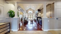 This expansive single-floor space is thoughtfully designed for both socializing and relaxation, combining elegant architectural details with versatile amenity areas. Stately white columns frame the open-plan interior, drawing the eye toward soaring vaulted ceilings accented by a chandelier and recessed lighting. Rich, glossy hardwood flooring flows throughout, creating warmth and visual continuity across each zone.

The main lounge area centers around a sleek tile-surround fireplace topped by a large wall-mounted television, flanked by French doors that invite abundant natural light and views of the surrounding outdoor landscape. Comfortable seating, including a leather sectional and accent chairs, creates an inviting atmosphere for gathering.

Adjacent to the lounge, a dedicated billiards room features a full-size pool table beneath a distinctive pendant light, wainscoting-paneled walls, and an additional wall-mounted television. The galley-style kitchen is well-appointed with dark cabinetry, granite countertops, stainless steel appliances, a stone tile backsplash, and pendant lighting. A large arched transom window at the far end of the main hall floods the space with natural light, completing the refined yet welcoming ambiance.