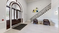 This sophisticated space makes an immediate impression with its grand foyer, where a soaring arched entryway framed by dark wood double doors and intricate leaded glass sets a refined tone. The expansive entry hall features soaring ceilings, crisp white walls accented by classical crown molding and wainscoting, and light-toned tile flooring that flows seamlessly throughout. A striking wrought-iron staircase with ornate scrollwork rises along one wall, complemented by a seating area with leather armchairs and contemporary artwork.

The open lobby area connects to multiple functional zones, including a well-appointed office space with large windows, elegant blue accent chairs, and a generous work desk set atop a patterned area rug. The fully equipped kitchen showcases white shaker cabinetry, granite countertops, a mosaic tile backsplash, stainless steel appliances, and a large center island with a built-in cooktop and professional-grade range hood. Natural light filters in through exterior-facing windows and French doors, offering views of lush greenery beyond.

A comfortable lounge area provides a welcoming retreat, anchored by a classic fireplace beneath a wall-mounted television, soft neutral furnishings, and tasteful decorative accents. Throughout the space, a consistent palette of whites, grays, and soft blues creates a polished, professional, and inviting atmosphere.