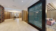 Located at 1715 US-46 E in Parsippany, NJ, this thoughtfully designed lobby space makes a strong first impression with its eclectic yet cohesive interior design. The expansive single-floor layout is defined by large-format light stone tile flooring in the main entry corridor, transitioning to warm wood-look plank flooring throughout the lounge and meeting areas.

A standout architectural feature is the oversized teal laser-cut decorative screen panel, framed in dark metal, which serves as a dramatic room divider between the entry corridor and the lounge area. Vertical wood slat wall panels appear throughout, adding texture and warmth. A circular recessed ceiling detail anchors the reception area, where a bold floral wallcovering in dark tones with vibrant pinks and greens adds a lively, artistic focal point alongside a decorative gold-accented console table.

The lounge area is furnished with a blush pink channel-tufted sofa, teal velvet accent chairs, and a marble-top round table, all set atop a bold zebra-print area rug. A sculptural glass globe chandelier illuminates the space above. Adjacent to the lounge, a glass-walled private meeting room features a marble round conference table, gold-accented task chairs, and a brass floral pendant light, offering a refined and functional space for gatherings.