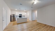 This single-floor residence offers a thoughtfully designed open-concept layout that seamlessly connects the kitchen, dining, and living areas. Light-toned hardwood-style flooring flows throughout the main living spaces, creating a bright and cohesive aesthetic. The kitchen serves as a central focal point, featuring crisp white shaker-style cabinetry, granite countertops with subtle gray and white tones, a tile backsplash, and a full suite of stainless steel appliances including a refrigerator, range, microwave, and dishwasher. A central island with a built-in sink provides additional prep space and serves as a natural gathering point. Stylish pendant lighting illuminates the island, complemented by a ceiling fan in the living area. A hallway leads to the private quarters, where carpeted bedrooms offer a comfortable retreat. The bedroom features a window that allows natural light to filter in. The bathroom is well-appointed with a granite-topped vanity, white cabinetry with brushed gold hardware, a large mirror, and a soaking tub. A generously sized walk-in closet with wire shelving provides ample storage. The overall design is clean, modern, and move-in ready.