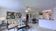 This single-floor residence offers an open-concept floor plan that seamlessly connects the living, dining, and kitchen areas, creating a spacious and cohesive environment ideal for everyday living and entertaining. The living room is generously sized, featuring plush carpeting, a ceiling fan with a light fixture that provides both comfort and illumination, and large decorative wall art that adds a contemporary touch to the space. Natural light flows throughout the main living area, enhancing the warm, neutral color palette of the walls and furnishings. The dining area sits adjacent to the living room and is defined by a pendant light overhead, providing an intimate setting for meals. The kitchen is positioned at the periphery of the open layout, featuring a raised breakfast bar, white cabinetry, and standard appliances including a full-size refrigerator, making it both functional and accessible. A hallway visible from the main living area leads to additional rooms, including a bathroom, offering a practical and well-organized layout. The consistent use of light tones across the carpet, walls, and fixtures gives the entire space a clean, airy ambiance that feels both inviting and easy to personalize.