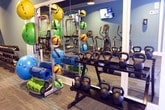 Located at 550 St. Michaels Way in Newport News, Virginia, this fitness center offers a well-appointed, single-floor exercise facility designed to accommodate a wide range of workout routines. The space features a striking interior palette of deep navy ceilings contrasted against cool blue-gray walls, accented with crisp white wainscoting that lends the room a clean, polished aesthetic. Durable, textured carpet tiles cover the workout floor throughout, providing comfort underfoot during training sessions.

The cardio zone is generously equipped with multiple treadmills, elliptical trainers, and upright stationary bikes, each positioned for easy access and personal space. Wall-mounted flat-screen televisions are strategically placed throughout the facility, keeping members entertained during workouts. Pendant and recessed lighting illuminate the space evenly, creating an energetic yet focused atmosphere.

The free weights area features a full rack of dumbbells, an adjustable workout bench, and a variety of stability and medicine balls for functional training. A dedicated strength and cable machine section rounds out the equipment offerings, supported by large mirrors that enhance the sense of openness and allow for form checks during exercise. Natural light filters in through windows along one wall, adding to the bright and inviting environment. The layout flows intuitively, making the space practical and welcoming for all fitness levels.