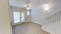 This two-story space is designed with a light, open aesthetic that creates a welcoming and functional environment. The main living area is finished with light hardwood-style flooring that runs throughout, complementing the crisp white walls and classic crown molding that frame the space with a polished touch. A ceiling fan with integrated lighting is centrally positioned, ensuring comfortable airflow and ambient illumination.

Natural light enters generously through a set of double French doors fitted with blinds, alongside an additional window above, brightening the room throughout the day. A wall-mounted sconce near the staircase provides supplementary accent lighting. The white-painted staircase with a classic baluster railing connects the main level to the upper floor, serving as both a functional and decorative architectural element.

Baseboards and trim throughout are finished in white, reinforcing the clean, cohesive design. The overall ambiance is neutral and move-in ready, offering a versatile canvas suited to a variety of interior styles. The layout provides a comfortable sense of openness while maintaining a warm, residential character across both levels.