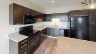 This single-floor residence showcases a contemporary design with wood-look flooring throughout, neutral tones, and an open layout that promotes a sense of flow and functionality. The kitchen serves as a focal point, featuring rich dark cabinetry, granite countertops, a central island with matching stone surface, and a full suite of black stainless steel appliances including a range, microwave, dishwasher, and refrigerator. A statement pendant light adds a striking decorative touch above the island. The open living area is bright and airy, benefiting from natural light that pours in through sliding glass doors leading to a private balcony with outdoor views of the surrounding landscape. The bedroom offers a comfortable and practical layout, complete with a dedicated walk-in closet outfitted with built-in shelving and hanging rods, along with an additional door providing flexible access. A large window fills the room with natural light. The bathroom is finished with clean, modern lines, featuring wood-look tile flooring, a full soaking tub with shower and curved curtain rod, a toilet, and a vanity topped with speckled granite countertops set in dark cabinetry. Together, the spaces create a cohesive, modern living environment that balances style and practicality.