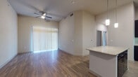 Located at 2223 Waterloo City Lane in Austin, Texas, this single-story residence offers a thoughtfully designed layout with a clean, contemporary aesthetic throughout. The open living area immediately captures attention with its notably high ceilings and warm wood-style flooring that reflects natural light filtering through large vertical-blind-covered sliding doors. A ceiling fan adds both function and character to the space, while a granite-topped kitchen island serves as a natural dividing point between the living and kitchen areas.

The kitchen is a standout feature, finished with bold dark cabinetry that contrasts sharply against a classic white subway tile backsplash. Stainless steel appliances—including a refrigerator with ice maker, range, and built-in microwave—are complemented by pendant lighting overhead and under-cabinet illumination, creating a polished, modern atmosphere. Light wood-tone flooring continues throughout the kitchen, maintaining visual cohesion.

The bedrooms offer plush carpeting and tall ceilings, with natural light provided through blinded windows. At least one bedroom includes access to a spacious walk-in closet with built-in shelving and hanging rods. The bathroom features a soaking tub with tiled surround and marble-look tile flooring, lending a clean, spa-like finish to the space.