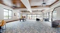 Located at 6135 S Highland Dr in Holladay, Utah, this single-floor community amenity space offers a generously sized, open-plan lounge designed for recreation and social gatherings. The room is anchored by a striking tray ceiling with warm wood paneling and recessed lighting, complemented by oversized ceiling fans that enhance the airy atmosphere throughout.

The space is defined by an abundance of natural light, courtesy of expansive floor-to-ceiling windows and wide sliding glass doors that line multiple walls. These openings provide seamless views and direct access to an outdoor courtyard and pool area, blurring the line between indoor and outdoor living.

A bold blue patterned carpet flows across the majority of the floor, creating a cohesive and vibrant foundation for the various activity zones. The game area features a full-size pool table and vintage arcade machines, while a putting green and a hanging hammock chair add additional recreational options. A lounge seating area with leather sofas and a coffee table is positioned near the panoramic windows overlooking the pool.

Transitioning toward the dining zone, hardwood flooring introduces a warmer aesthetic. A round dining table with upholstered chairs sits beside a functional wet bar with cabinetry, a sink, and a wine rack — ideal for entertaining. A teal accent wall with decorative artwork adds a pop of color, tying the design together with a modern, welcoming character.