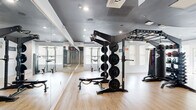 Located at 927 W Morgan St in Raleigh, NC, this fitness center offers a well-appointed exercise space designed to support a full range of workout routines. The single-floor layout provides an open, functional environment finished with light hardwood-style flooring, a dark painted ceiling, and recessed lighting that creates a modern, energetic atmosphere. Large windows with white shutters line the walls, allowing natural light to brighten the space throughout the day.

The exercise room is comprehensively outfitted for both strength and cardio training. A large multi-functional rig serves as the centerpiece, featuring a plate-loaded weight tree, pull-up bars, and various attachment points for functional fitness exercises. Accompanying this is a full dumbbell rack, a kettlebell rack, medicine balls, resistance bands, and a TKO heavy punching bag. Cardio equipment includes stationary bikes, a stair climber, and a treadmill. Floor-to-ceiling mirrors along one wall enhance the sense of space and allow for form monitoring during workouts.

A distinct accent wall featuring a dark hexagonal tile pattern adds a bold design element, while a warm wood-paneled wall near a lounge nook introduces texture and contrast. A water fountain is also available within the space, rounding out the amenities in this thoughtfully designed community fitness center.