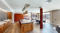 This sophisticated single-floor amenity lounge is designed with a seamless open-plan layout that connects a fully equipped kitchen, multiple lounge areas, and direct access to an outdoor terrace. The kitchen anchors the space with a substantial island topped in dramatic dark veined stone, warm wood cabinetry, a textured metallic tile backsplash, and a row of cascading brass pendant lights that serve as a striking focal point overhead. Rich hardwood flooring flows throughout, unifying each zone of the space.

The lounge areas are defined by bold terracotta-toned wainscoted walls accented with decorative panel molding and ambient cove lighting. Seating options range from a generous sectional sofa to a curated arrangement of a leather sofa and coral accent chairs around a marble-topped coffee table, offering both casual and social settings. A stone-surround fireplace with a wall-mounted display screen provides a warm gathering point.

Floor-to-ceiling glass walls and sliding doors flood the interior with abundant natural light and open directly onto a furnished outdoor terrace, complete with pergola structures and additional seating. Deep-toned structural columns add an architectural grounding element throughout, while the careful layering of materials and color creates an inviting, upscale ambiance suited for entertaining and relaxation alike.