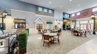 Located on Amity Drive in Fort Wayne, Indiana, this single-floor amenity center offers a thoughtfully designed community space that creatively replicates a small-town streetscape entirely indoors. The centerpiece is a soaring, open town square concept with dramatic double-height ceilings, upper clerestory windows, and themed storefronts that include a coffee shop, Monty's Pub, a hair salon, and a post office — all rendered with authentic architectural details such as brick facades, awnings, stone accents, and decorative street lamps. The main gathering area is generously proportioned, furnished with round and square dining tables and wooden chairs arranged throughout a patterned carpet, ideal for socializing and community dining. Adjacent lounge areas offer comfortable, residential-style seating with neutral-toned sofas, plaid accent chairs, and warm table lamps, accented by framed artwork and natural greenery. The post office corridor features warm hardwood flooring, a full wall of resident mailboxes, and ample natural light from exterior-facing windows. Additional sitting rooms provide intimate, light-filled retreats with large windows dressed in sheer curtains and tasteful décor. The overall ambiance balances a welcoming, small-town charm with a clean, modern interior aesthetic throughout.
