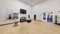Located at 401 Granby St in Norfolk, Virginia, this fitness center offers a comprehensive workout facility spread across a single floor with two distinct exercise zones. The cardio and strength area features vibrant, color-blocked accent walls in blue, green, red, and yellow, adding an energetic atmosphere to the white-walled space. This zone is equipped with treadmills, elliptical trainers, stationary bikes, a stair climber, a cable machine, and a rowing machine, complemented by a rack of dumbbells and kettlebells, a weight bench, and a wall-mounted flat-screen television. Large mirrors extend across one wall, enhancing the sense of space and allowing users to monitor their form. The second area transitions to a light hardwood floor, creating a dedicated functional training space. This room houses a multi-station cable and functional trainer system along with open floor space ideal for stretching, yoga, or floor exercises. Recessed ceiling lighting illuminates both areas throughout, while floor fans provide additional ventilation. French glass doors connect the two zones, ensuring a seamless flow through the facility. The overall design is clean, modern, and purposefully arranged to support a full range of fitness activities.