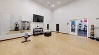 Located at 401 Granby St in Norfolk, Virginia, this fitness center offers a comprehensive workout facility spread across a single floor with two distinct exercise zones. The cardio and strength area features vibrant, color-blocked accent walls in blue, green, red, and yellow, adding an energetic atmosphere to the white-walled space. This zone is equipped with treadmills, elliptical trainers, stationary bikes, a stair climber, a cable machine, and a rowing machine, complemented by a rack of dumbbells and kettlebells, a weight bench, and a wall-mounted flat-screen television. Large mirrors extend across one wall, enhancing the sense of space and allowing users to monitor their form. The second area transitions to a light hardwood floor, creating a dedicated functional training space. This room houses a multi-station cable and functional trainer system along with open floor space ideal for stretching, yoga, or floor exercises. Recessed ceiling lighting illuminates both areas throughout, while floor fans provide additional ventilation. French glass doors connect the two zones, ensuring a seamless flow through the facility. The overall design is clean, modern, and purposefully arranged to support a full range of fitness activities.