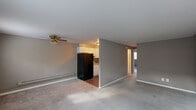 This single-floor unit is designed with a practical, open layout that connects the living and sleeping areas seamlessly. The living room serves as the central gathering space, featuring neutral gray-toned walls, plush wall-to-wall carpeting, and a ceiling fan with a brass finish. Large windows allow natural light to filter in, casting bright patches across the floor. Baseboard heating units run along the walls in multiple rooms, ensuring efficient warmth throughout. The front entry opens directly into the main living area, which includes a closet and an additional window for added brightness.

The kitchen is tucked adjacent to the living space and features white cabinetry, a black refrigerator, and wood-style flooring that contrasts with the carpeted areas. A pass-through opening connects the kitchen visually to the main living area, enhancing the sense of flow.

The bedrooms are carpeted and generously proportioned, each with windows overlooking greenery outside. One bedroom includes a window-mounted air conditioning unit alongside baseboard heating, offering year-round climate control. A bedroom with a bi-fold closet provides practical storage. The overall palette of warm gray and white tones lends the unit a clean, understated ambiance throughout.