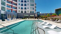 Located at 100 Pine St W in Wilson, North Carolina, this outdoor amenity space is a thoughtfully designed courtyard retreat set within a contemporary multi-story building complex. The centerpiece is a generously sized swimming pool with crystal-clear water, accessible via wide entry steps and stainless steel handrails, surrounded by an expansive concrete pool deck finished in a light aggregate surface. The sundeck is well-appointed with rows of modern white chaise lounges, offering ample space for relaxation under open skies. Scattered throughout the deck are shaded seating areas outfitted with red umbrellas and outdoor dining tables, providing comfortable spots for gathering. A dedicated lounge zone features modular white sectional furniture arranged on the deck, ideal for socializing. The courtyard also includes an outdoor bar-height table with bold red stools, set against a backdrop of lush, layered landscaping featuring young trees, ornamental shrubs, and brick retaining walls that add warmth and texture to the space. Manicured planting beds with mulched borders and stairway access points connect various levels of the outdoor area seamlessly. The surrounding multi-story building facade, rendered in a modern palette of white panels, gray siding, and red accent columns with private balconies, frames the courtyard with a striking architectural presence.