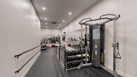 This fitness center in Boston, MA, is a thoughtfully designed single-room exercise facility that maximizes its compact footprint to deliver a comprehensive workout environment. The space features a clean, modern aesthetic with crisp white walls, recessed ceiling lighting, and dark rubberized flooring that provides both durability and traction during exercise.

Full-length mirrors line the walls, visually expanding the space and allowing users to monitor their form throughout workouts. The cardio section is equipped with a stationary spin bike featuring a mounted display screen, an elliptical trainer, and a treadmill. On the strength training side, a cable pulley machine offers versatile resistance training options, while a dedicated free weights area includes a rack of dumbbells in graduated sizes, a flat workout bench, and stability accessories such as medicine balls and balance balls.

Wall-mounted safety features, including an AED unit and fire extinguishers, are clearly positioned throughout the room, reflecting a strong emphasis on user safety. A ceiling-mounted HVAC unit ensures proper climate control and ventilation. Handrails along a central dividing section further enhance safety and accessibility. The overall ambiance is clean, functional, and professional, making this fitness center a well-equipped space for a full range of cardiovascular and strength training activities.