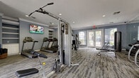 This fitness center in Houston, Texas, offers a comprehensive workout environment designed to accommodate a wide range of exercise routines. The space is finished with cool blue-gray walls, crown molding, and textured multi-tone carpet flooring, creating a clean and motivating atmosphere throughout.

The cardio section is anchored by a row of treadmills mounted along one wall, accompanied by elliptical machines and a stair climber, providing ample options for aerobic training. Strength training is well supported by a multi-station cable machine, a functional trainer, adjustable benches, and a dedicated rack of medicine balls for dynamic exercises. A water station and storage cabinetry are conveniently positioned to support users during their workouts.

Natural light fills the room through multiple arched and French-style windows and dual sets of glass-paneled doors that open to the outdoors, offering views of surrounding greenery. Recessed lighting supplements the natural light, ensuring the space remains well-lit at all times. A wall-mounted flat-screen television adds an entertainment element, particularly suited to those using the cardio equipment.

The layout is thoughtfully organized to allow users to move freely between stations without congestion, making it practical for simultaneous use. Overall, this fitness center delivers a fully functional and inviting environment for residents seeking a convenient on-site workout facility.