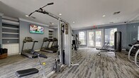 This fitness center in Houston, Texas, offers a comprehensive workout environment designed to accommodate a wide range of exercise routines. The space is finished with cool blue-gray walls, crown molding, and textured multi-tone carpet flooring, creating a clean and motivating atmosphere throughout.

The cardio section is anchored by a row of treadmills mounted along one wall, accompanied by elliptical machines and a stair climber, providing ample options for aerobic training. Strength training is well supported by a multi-station cable machine, a functional trainer, adjustable benches, and a dedicated rack of medicine balls for dynamic exercises. A water station and storage cabinetry are conveniently positioned to support users during their workouts.

Natural light fills the room through multiple arched and French-style windows and dual sets of glass-paneled doors that open to the outdoors, offering views of surrounding greenery. Recessed lighting supplements the natural light, ensuring the space remains well-lit at all times. A wall-mounted flat-screen television adds an entertainment element, particularly suited to those using the cardio equipment.

The layout is thoughtfully organized to allow users to move freely between stations without congestion, making it practical for simultaneous use. Overall, this fitness center delivers a fully functional and inviting environment for residents seeking a convenient on-site workout facility.