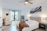 Located at Vintage Commerce in Fort Myers, Florida, this contemporary studio unit offers a thoughtfully designed single-level layout that maximizes both comfort and functionality. The open-concept main living area seamlessly integrates the sleeping, lounging, and dining zones, all finished with light wood-toned flooring that contributes to a bright, airy atmosphere throughout. A ceiling fan overhead provides comfort and enhances the spacious feel of the room. Large floor-to-ceiling windows and a glass-panel balcony door flood the space with abundant natural light while offering views of the surrounding community. The balcony provides a pleasant outdoor retreat. The kitchen is a standout feature, showcasing sleek dark cabinetry with gold hardware, crisp white countertops, and a striking teal glazed subway tile backsplash that adds a bold pop of color. Stainless steel appliances, including a refrigerator, range, and over-the-range microwave, complete the modern culinary space. The clean white walls and neutral palette throughout the studio create a cohesive, contemporary aesthetic, making the space feel polished and move-in ready. This studio is ideal for those seeking a modern, low-maintenance living space in Fort Myers.