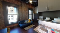 Located in the heart of downtown Chattanooga at 818 Georgia Ave, this urban studio unit showcases a compelling blend of historic industrial character and modern design. The open-concept living area is anchored by striking exposed brick walls, dark hardwood-style flooring, and dramatic ceiling heights that lend an expansive, loft-like feel throughout. An exposed ceiling with visible ductwork and industrial piping reinforces the raw architectural aesthetic, while a ceiling fan and ductless mini-split HVAC unit ensure year-round comfort.

Tall, black-framed windows flood the living space with natural light and frame views of Chattanooga's surrounding historic architecture. The living area accommodates a sofa, dining table, and seating, creating a well-defined yet flexible layout. A built-in kitchenette provides cabinet storage, a countertop, and a sink for everyday convenience.

The sleeping area is partitioned within the open floor plan, featuring a platform bed with integrated storage drawers and an additional kitchenette unit with overhead cabinetry. The private bathroom offers a clean, modern finish with tile flooring, a dark vanity with mirror, and a toilet — completing this efficient, thoughtfully designed urban living space.