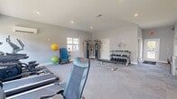 This fitness center is a thoughtfully designed single-floor facility comprising a generously sized exercise room and a dedicated bathroom. The exercise room is bright and open, featuring light gray walls, recessed ceiling lighting, and wall-to-wall carpet flooring that provides a comfortable surface underfoot. Multiple windows allow natural light to fill the space, contributing to an energizing workout atmosphere, while a wall-mounted split-unit air conditioner ensures year-round climate comfort.

The exercise room is comprehensively equipped to support a wide variety of workouts. Cardio equipment includes treadmills and an elliptical machine, while a multi-functional cable machine provides options for a full range of strength training exercises. A three-tier dumbbell rack stocked with a complete set of free weights, a weight bench, and an adjustable incline bench cater to resistance training needs. Functional training accessories such as medicine balls, stability balls, and resistance tools are neatly organized on a dedicated rack. A motivational "Workout" wall decal adds an inspiring finishing touch to the space.

The facility is accessed through glass-paneled double doors. The bathroom is clean and modern, featuring a floating white vanity with a rectangular sink, brushed nickel fixtures, a large wall mirror, and warm wood-look flooring, providing a polished and convenient post-workout amenity.