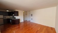 This single-floor unit is defined by rich, warm-toned hardwood flooring that runs continuously throughout the main living spaces, creating a cohesive and inviting atmosphere. The open-concept layout connects the living area to the kitchen, maximizing the sense of space and promoting an efficient flow between functional zones.

The kitchen is thoughtfully designed with dark espresso cabinetry extending to the ceiling, providing ample storage. Light granite countertops offer a striking contrast to the cabinetry, while stainless steel appliances — including a gas range, refrigerator, and built-in microwave — complete the functional setup. A ceiling-mounted light fixture illuminates the workspace effectively.

The living area is generously sized, featuring clean white walls and a large window that allows substantial natural light to fill the room. A wall-mounted air conditioning unit ensures year-round climate comfort. The open sightlines from the living area to the kitchen enhance the overall spaciousness of the unit.

The bedroom is a well-proportioned, private space continuing the hardwood flooring and white walls, with a large window providing additional natural light and exterior views.

The bathroom features a clean, modern design with a white basin sink, a combination tub and shower, and decorative striped gray tile accenting the shower surround, complemented by neutral mosaic tile on the back wall.