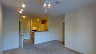 This single-floor residential unit offers a practical and open layout designed for comfortable everyday living. The living area is finished with soft wall-to-wall carpeting, creating a warm and inviting atmosphere, while a ceiling-mounted multi-bulb light fixture serves as a functional centerpiece overhead. Track lighting is installed throughout, providing adjustable, well-distributed illumination across the space.

The kitchen is situated in an open-concept arrangement, separated from the living area by a white peninsula counter finished with a dark countertop surface. This design choice allows the kitchen to feel connected to the main living space while maintaining a defined cooking zone. The kitchen features light wood cabinetry, under-cabinet lighting, and includes standard appliances such as a range and refrigerator. The kitchen flooring transitions to a tiled surface, clearly delineating the two areas.

Multiple doorways are visible throughout the unit, suggesting access to additional rooms such as bedrooms or a bathroom. The walls are finished in a clean, neutral white tone, contributing to a bright and airy feel. The layout is efficient and well-organized, making thoughtful use of the available space. Overall, the unit presents a functional and straightforward residential environment suited for a range of occupants.