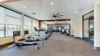 This expansive fitness center offers a comprehensive workout environment thoughtfully designed across a single open floor. The layout is divided into distinct functional zones, creating an organized and efficient training experience. The cardio zone lines one side of the space, featuring a full row of treadmills, ellipticals, rowing machines, and stationary bikes, all positioned to take advantage of large windows fitted with roller shades that allow natural light to filter throughout the room.

The strength training area occupies the opposite side and is anchored by an extensive dumbbell rack with a full weight range, an adjustable bench, a cable machine, and a dedicated functional training zone equipped with battle ropes and additional resistance equipment. Floor-to-ceiling mirrors along the strength zone wall visually expand the space and provide exercise feedback.

Architectural details include warm wood-toned accent beams that frame transitions between zones, white walls, and neutral carpet tile flooring throughout. Oversized black ceiling fans contribute to air circulation. A glass-paned door along one wall provides direct access to the outdoor courtyard, where palm trees and a landscaped patio are visible. A separate enclosed room with glass doors houses cycling equipment. The overall ambiance is modern, clean, and professionally outfitted.