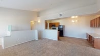 This single-floor residence features an open-concept design that integrates the living and kitchen areas into one cohesive space. The living area is finished with plush, neutral-toned carpeting that adds warmth underfoot, while the kitchen and adjoining areas transition to hardwood-style flooring for easy maintenance.

The kitchen is defined by a built-in peninsula that separates it from the living space, offering both a functional prep surface and a casual dining or entertaining perch. Warm medium-toned wood cabinetry lines the upper kitchen walls, providing ample storage, while black appliances — including a refrigerator and microwave — add a contemporary contrast. A ceiling-mounted vent ensures proper ventilation throughout the cooking space.

The ceilings are notably elevated, contributing to an airy, spacious feel throughout the interior. Subtle wall sconces provide warm accent lighting in key areas, complementing the neutral, light-colored walls that keep the space feeling bright and welcoming.

A balcony extends the living space outdoors, offering views of the surrounding residential community, covered parking structures, and open skies. The exterior setting features mature landscaping and a well-maintained community environment, visible from this elevated vantage point.
