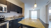 This single-floor space showcases a seamlessly connected kitchen and living area, creating an open and functional layout. The kitchen is appointed with rich, dark cabinetry paired with a classic white subway tile backsplash, offering a striking contrast that anchors the space. A large granite island with warm gold and cream tones takes center stage, providing generous prep space and serving as a natural gathering point. A pendant light with a brass finish hangs above the island, adding a refined decorative touch. The kitchen also features a double stainless steel sink, matching dark lower cabinetry, and a stainless steel dishwasher, combining practicality with a cohesive aesthetic.

The space transitions effortlessly into the living area, where a ceiling fan with an integrated light fixture provides both airflow and illumination. A full-length sliding glass door with vertical blinds floods the area with natural light and offers direct access to the outdoors. The combination of tile flooring, neutral walls, and thoughtful lighting throughout creates a bright, modern, and welcoming ambiance well-suited to everyday living.