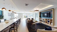 Located at 500 Broadway in Malden, MA, this single-floor amenity space offers a thoughtfully designed environment for socializing and recreation. The open-plan layout is anchored by a generously sized lounge area featuring plush seating arrangements, a large wall-mounted television set within a custom built-in shelving unit, and a dark area rug that defines the space against warm light wood flooring found throughout.

A long bar-height counter with seating runs along one side of the lounge, ideal for casual gatherings. Adjacent to it, a well-appointed wet bar area features sleek cabinetry, white quartz countertops, an integrated sink, and under-counter appliances, all set against a bold teal accent wall. Decorative pendant lighting with warm-toned wood accents illuminates both the bar and lounge zones, while cove lighting adds a refined finishing touch to the tray ceilings.

Large windows and glass doors flood the space with natural light and offer views of an outdoor patio area featuring a pergola and stone fireplace. A separate game corridor, distinguished by a striking geometric blue ceiling installation, provides a shuffleboard table and additional lounge seating. The overall design palette of soft grays, navy, and warm wood tones creates a welcoming, contemporary ambiance throughout.