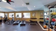 The fitness center is a dedicated single-floor workout space designed to support a well-rounded exercise routine. The room features warm yellow walls paired with a light ceiling accented by recessed lighting, creating a bright and energizing atmosphere. Large windows along one wall allow natural light to filter in through slatted blinds, contributing to an open, airy feel. The flooring is covered in durable rubber matting, providing cushioning and traction suitable for a variety of workouts. A full-length mirror runs along one wall, visually expanding the space while allowing users to monitor their form during exercise. The cardio section includes two treadmills and an elliptical machine, all equipped with digital displays. The free weight area features a tiered dumbbell rack stocked with a wide range of weights, accompanied by an adjustable workout bench. Additional weight storage racks are positioned nearby for convenient access. Decorative framed artwork with a nautical theme adds a subtle finishing touch to the walls, balancing the functional nature of the space with a touch of character. Ceiling ventilation ensures adequate airflow throughout workouts.