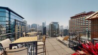 Located at 100 W Chestnut St in Chicago's vibrant Near North Side neighborhood, this rooftop sun deck at West Chestnut Apartments delivers a compelling outdoor retreat high above the city streetscape. The deck is accessed via a staircase framed by classic brick masonry, with sturdy metal railings guiding residents up to the open-air level. Once on the deck, the expansive terrace is finished with a durable aggregate tile surface and enclosed by sleek steel railings that preserve unobstructed sightlines in every direction. The space is thoughtfully furnished with high-top wooden dining tables and chairs ideal for casual gatherings, as well as reclining lounge chairs perfectly positioned for sunbathing and relaxation. Decorative planters with seasonal flowers add a touch of warmth to the urban setting. The defining feature of this rooftop is its sweeping, panoramic view of the Chicago skyline, with prominent downtown towers — including iconic skyscrapers — rising dramatically against the open sky. The sun deck operates daily from 8AM to 11PM and accommodates up to 27 guests, making it a shared community amenity suited for both quiet relaxation and social occasions. Signage from West Chestnut Apartments outlines guidelines to ensure an enjoyable experience for all residents.