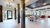 Located at 450 S State St in Clearfield, Utah, this thoughtfully designed single-floor amenity space offers a generous and versatile layout ideal for gathering and recreation. Light wood-look flooring flows throughout, complementing the contemporary aesthetic defined by tray ceilings with dark wood accents, recessed lighting, and stylish pendant and chandelier fixtures.

The open kitchen is a standout feature, equipped with navy cabinetry, a striking chevron-patterned tile backsplash, stainless steel appliances including a full-size refrigerator, a stainless range hood, and a large white quartz island with bar seating — perfect for casual dining or catered events.

The living area is warmly appointed with a mix of seating options, including plush leather sofas, bold orange barrel chairs, and upholstered booth-style banquettes with wood tables. Large windows invite abundant natural light and offer views of the surrounding exterior. A dedicated lounge nook includes a large flat-screen TV, making it ideal for relaxed viewing.

A separate game room features a full-size billiards table, built-in bench seating, and decorative wall sconces beneath a dramatic dark wood ceiling. Dark wood columns frame the main entry, which opens via glass double doors to the outdoors. The overall ambiance is modern, welcoming, and well-appointed throughout.