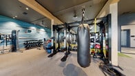 This modern fitness center is thoughtfully designed to support a full range of workout disciplines across a single, expansive floor. The space is divided into distinct functional zones, each outfitted with professional-grade equipment. The cardio area features multiple treadmills, elliptical machines, and a rowing machine positioned in front of large floor-to-ceiling windows that flood the space with natural light and offer views of the surrounding outdoor area. The strength training zone is anchored by a comprehensive dumbbell rack, cable machines, and adjustable benches, all set against a vivid blue abstract mural that serves as a dramatic focal point. A dedicated functional training area includes ceiling-mounted heavy bags, Adidas-branded multi-functional training rigs, TRX suspension systems, turf flooring for sled or agility work, medicine balls, and an assortment of specialty fitness accessories. Structural columns finished in warm wood tones contrast with the dark ceiling and charcoal walls, giving the space a sleek, modern aesthetic. Full-length mirrors are strategically placed throughout to enhance the sense of openness. Additional amenities include a water fountain station set into a tiled accent wall. The overall ambiance is energetic yet refined, reflecting a high-end facility designed to meet diverse fitness needs.