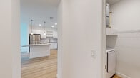 This single-floor space presents a cohesive, contemporary design highlighted by light hardwood flooring and a crisp white color palette that runs throughout. The kitchen serves as a central feature, offering white flat-panel cabinetry extending to the ceiling, a classic subway tile backsplash, and granite countertops across both the perimeter workspace and a central island. Stainless steel appliances, including a refrigerator, range, and microwave, contribute to a polished, functional aesthetic. Pendant lighting above the island and recessed ceiling lights provide layered illumination.

The living and bedroom areas are bright and open, with large windows allowing natural light to fill the space. A ceiling fan in the primary bedroom adds practical comfort. The bathroom is well-appointed with a granite-topped vanity, white cabinetry, and matching hardware, maintaining the consistent modern aesthetic. A spacious walk-in closet features multi-level wire shelving for organized storage. The dedicated laundry area houses a full-size washer and dryer alongside a water heater, adding everyday convenience. Overall, the space presents a clean, modern, and functional environment.