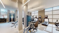 This fitness center offers a thoughtfully designed, single-floor layout that accommodates a full spectrum of workout needs. The space features a clean, contemporary aesthetic with neutral warm tones, light wood-style flooring, and a combination of recessed and linear LED lighting that ensures even illumination throughout.

Large floor-to-ceiling windows line the perimeter, flooding the space with natural light and providing views of the surrounding neighborhood. Full-length mirrors are strategically placed along the walls, visually expanding the space and allowing users to monitor their form during exercise.

The cardio zone is well-appointed with multiple treadmills and elliptical machines, each equipped with individual digital displays. A rowing machine and stationary bike add further variety to the cardio offerings. The strength training area features a robust selection of selectorized cable machines and multi-functional units suited for a wide range of resistance exercises.

The free weights section includes a comprehensive dumbbell rack spanning various weight increments, along with a barbell and weight plate station. A dedicated functional training zone is stocked with medicine balls, battle ropes, balance trainers, and plyometric boxes, catering to dynamic and sport-specific training.

The overall ambiance is professional, clean, and inviting, reflecting a high-quality facility designed to support diverse fitness goals.
