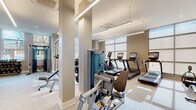 This fitness center offers a thoughtfully designed, single-floor layout that accommodates a full spectrum of workout needs. The space features a clean, contemporary aesthetic with neutral warm tones, light wood-style flooring, and a combination of recessed and linear LED lighting that ensures even illumination throughout.

Large floor-to-ceiling windows line the perimeter, flooding the space with natural light and providing views of the surrounding neighborhood. Full-length mirrors are strategically placed along the walls, visually expanding the space and allowing users to monitor their form during exercise.

The cardio zone is well-appointed with multiple treadmills and elliptical machines, each equipped with individual digital displays. A rowing machine and stationary bike add further variety to the cardio offerings. The strength training area features a robust selection of selectorized cable machines and multi-functional units suited for a wide range of resistance exercises.

The free weights section includes a comprehensive dumbbell rack spanning various weight increments, along with a barbell and weight plate station. A dedicated functional training zone is stocked with medicine balls, battle ropes, balance trainers, and plyometric boxes, catering to dynamic and sport-specific training.

The overall ambiance is professional, clean, and inviting, reflecting a high-quality facility designed to support diverse fitness goals.