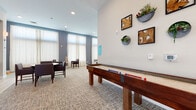 This well-appointed community amenity floor offers a thoughtfully designed collection of social and recreational spaces. The bright and airy lounge area serves as a central gathering point, featuring plush seating including wingback chairs and a cream sofa, all set beneath soaring ceilings and bathed in natural light from floor-to-ceiling windows dressed with sheer white curtains. French doors lead to an outdoor balcony, further enhancing the open, connected feel of the space. A cozy library nook showcases a built-in teal bookshelf stocked with books and décor, complemented by leather wingback chairs and round side tables — ideal for quiet reading or casual conversation. The game room is a standout feature, housing both a full-size shuffleboard table and a pool table, alongside a large wall-mounted TV set against a stacked stone accent wall with a built-in bar and beverage refrigerators. A golf simulator adds another layer of entertainment. Smaller dining and meeting areas are thoughtfully placed throughout, including a bistro-style table near a sliding barn wood door and additional seating near panoramic windows with outdoor views. Neutral carpeting, curated wall art, and live greenery accents tie the entire space together with a warm, welcoming ambiance.