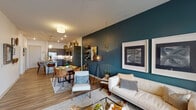 Located at 6004 Grandover Village Rd in Greensboro, NC, this single-floor residence offers a thoughtfully designed open-concept layout that seamlessly connects the living, dining, and kitchen areas. The living room makes a striking impression with a bold teal accent wall, large windows that invite natural light, a ceiling fan, and stylish mid-century modern furnishings. The kitchen is a standout feature, equipped with gray shaker-style cabinetry, dark granite countertops, a white subway tile backsplash, stainless steel appliances, and a center island with bar seating — all beneath elegant pendant lighting. Wood-look flooring flows throughout the main living areas, adding warmth and cohesion to the space. The bedroom offers a cozy retreat highlighted by a rich amber accent wall, plush carpeting, a ceiling fan, and generous natural light from a side window. The bathroom features a dual-sink vanity with dark cabinetry and light countertops, tile flooring, and a full soaking tub. A dedicated in-unit laundry area with a full-size washer and dryer adds everyday convenience. Throughout the residence, carefully chosen lighting fixtures and décor elements contribute to a modern yet inviting ambiance.