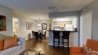 This single-floor residence offers a thoughtfully designed open-concept layout that effortlessly connects the main living areas. The kitchen features light wood cabinetry, dark countertops, and white appliances, and opens to the rest of the living space via a raised bar counter fitted with sleek black bar stools — ideal for casual dining or entertaining. The dining area sits adjacent to the kitchen, accommodating a round table with cushioned dark wood chairs beneath a classic chandelier that adds a warm, inviting glow. Framed artwork on the neutral-toned walls adds a touch of personality and character to the space. Rich hardwood flooring flows throughout, anchoring the open layout and providing a warm contrast to the light-colored walls and ceiling. The living area extends naturally from the dining space, with glimpses of comfortable seating visible at the edges of the room, reinforcing the cohesive and functional flow of the floor plan. Recessed lighting and pendant fixtures provide layered illumination throughout, creating a bright and welcoming ambiance across the entire space.