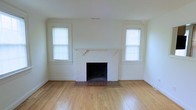 This single-floor space offers a warm and inviting atmosphere rooted in classic architectural details. The main living area is anchored by a white-painted brick fireplace, which serves as a natural focal point and adds timeless character to the room. Flanking the fireplace are two symmetrically placed windows dressed with horizontal blinds, allowing an abundance of natural light to flow throughout the space and creating a bright, airy feel. The floors throughout are finished in traditional hardwood, lending warmth and durability to the interior while complementing the clean, neutral color palette. The walls and trim are painted in a crisp white, reinforcing a sense of openness and making the space feel fresh and versatile. The ceiling is smooth and unadorned, maintaining a clean aesthetic that keeps the focus on the room's architectural features. The overall ambiance is understated and classic, offering a solid foundation that is well-suited to a variety of interior styles. The layout and design reflect a straightforward, functional approach while retaining the charm of traditional construction details such as the fireplace surround and the original hardwood flooring.