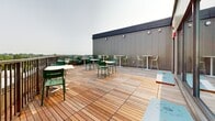 Located at 1190 Hancock St in Quincy, Massachusetts, this rooftop terrace offers a well-appointed outdoor amenity space with expansive open-air views of the surrounding Quincy skyline and neighboring greenery. The terrace features a warm-toned hardwood deck that extends generously across the rooftop, creating an inviting and versatile gathering area suitable for both casual relaxation and social events.

Seating is thoughtfully arranged throughout the space, combining standard-height white tables paired with green slatted chairs and elevated bistro-style high-top tables with light-colored bar stools. This mix of seating options accommodates a variety of uses, from informal lounging to more structured social interaction. A cornhole board is also present, adding a recreational element to the space.

Large sliding glass doors connect the terrace to an adjacent interior area, enhancing the flow between indoor and outdoor environments while allowing for natural light and cross ventilation. The perimeter of the deck is lined with a railing system that ensures safety while preserving the open sightlines to the cityscape and sky beyond.

The overall design aesthetic is clean and contemporary, with neutral-toned panel cladding along the building façade contrasting against the richness of the natural wood decking. The space is well-suited for community gatherings, offering a functional and visually appealing outdoor retreat above the streets of Quincy.