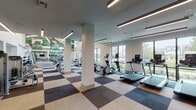 This fitness center in Houston, TX (77008) is a thoughtfully designed, single-floor facility that offers a comprehensive workout environment across multiple interconnected zones. The space is characterized by its striking multi-tone checkered carpet flooring in shades of navy, taupe, beige, and charcoal, which visually defines each area while maintaining a cohesive aesthetic throughout.

The cardio zone features a row of treadmills and elliptical machines positioned directly in front of large floor-to-ceiling windows, providing views of the surrounding outdoor greenery and encouraging an energizing workout experience. A dedicated strength training area houses a full selection of cable machines, weight benches, and a dumbbell rack, all illuminated by sleek linear pendant lighting.

A functional training zone is equipped with a heavy punching bag, TRX suspension systems, and battle rope anchors, catering to high-intensity training needs. An open studio area with ceiling fans, recessed lighting, and mirrored walls offers ample space for stretching, yoga, or group fitness activities, with stability balls and accessories stored nearby.

Soaring double-height ceilings in the main workout area, combined with expansive windows on multiple walls, flood the space with natural light and create an open, airy ambiance. White structural columns and neutral wall tones contribute to the clean, modern design of this well-appointed fitness facility.