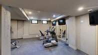 Located at 525 Main St in Hartford, CT, this fitness center offers a well-equipped exercise space designed to support a range of workout routines. The room is finished with durable, textured carpet flooring throughout and is illuminated by recessed ceiling lighting, creating a functional and comfortable environment for physical activity.

The cardio zone is anchored by True-brand equipment, including elliptical trainers, a recumbent bike, and a rowing machine. A framed wall-mirror installation visually expands the space and allows users to monitor their form during workouts. Multiple wall-mounted flat-screen televisions are positioned throughout the room for entertainment during exercise sessions.

A dedicated strength training area features a tiered dumbbell rack stocked with free weights, a adjustable weight bench, and additional strength equipment. A built-in bench along one wall provides a convenient seating area, positioned beneath a pair of small hopper windows that allow in natural light. A structural support column divides the room into distinct functional zones without significantly disrupting the open flow of the space.

The neutral-toned walls and low-profile ceiling give the room a clean, utilitarian aesthetic. Ventilation is provided via ceiling-mounted HVAC registers, and safety is addressed with visible smoke detectors and emergency exit signage. Overall, this fitness center delivers a practical and organized workout environment.