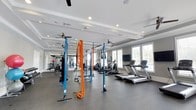 This modern fitness center is designed to accommodate a wide range of workout routines within a single, open floor layout. The space features elegant tray ceilings with recessed lighting and multiple ceiling fans, creating a bright and comfortable environment for exercise. Large windows along one wall allow generous natural light to fill the room, offering views of the outdoors and contributing to an airy, energizing atmosphere. The flooring consists of durable rubber gym matting, providing both safety and functionality throughout the space. Equipment is thoughtfully arranged to maximize usability, and includes a row of cardio machines such as treadmills and ellipticals positioned near the windows, as well as a multi-station strength training rig with TRX suspension systems and battle ropes for functional fitness training. Additional strength and cable machines are visible toward the rear of the space, offering options for a comprehensive full-body workout. A wall-mounted flat-screen television is positioned for easy viewing during cardio sessions. The overall ambiance is clean, professional, and well-maintained, reflecting a facility designed to support a variety of fitness goals in a spacious and well-lit setting.