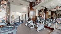 Located at 3850 W Northwest Highway in Dallas, TX, this well-appointed fitness center offers a comprehensive and energizing workout environment. The space features soaring ceilings that contribute to an open, airy atmosphere, complemented by expansive floor-to-ceiling windows that flood the interior with natural light and provide views of the surrounding landscaped greenery.

The facility is outfitted with a robust selection of Life Fitness commercial-grade equipment, including multiple treadmills, elliptical trainers, stationary bikes, and a cable machine, catering to both cardio and strength training needs. A dedicated free weights area features an organized dumbbell rack alongside adjustable benches, supporting a full range of strength workouts.

Design elements elevate the space beyond a standard gym. Large-format motivational photo murals depicting athletes in action span multiple walls, creating an inspiring visual backdrop. Full-length mirrored wall panels with rich dark wood accents add depth to the room while allowing users to monitor their form. The flooring transitions between durable rubber and patterned carpet tiles, clearly delineating functional zones throughout the space. Recessed lighting throughout ensures bright, even illumination across the entire floor, making this fitness center a well-designed and fully equipped destination for residents and users alike.